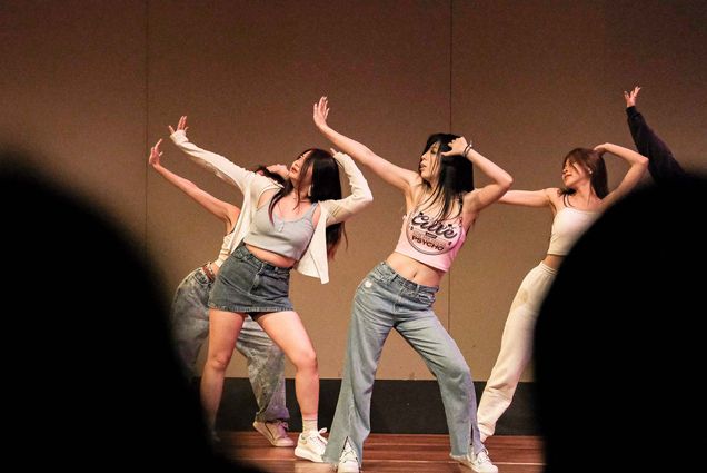 Photo of Students from G-Power, a BU K-Pop Dance Company presenting their hip-pop dance during the Student Showcase at Tsai Performance Center. A group of AAPI women on stage raise their hands over their heads to their right; they wear jeans, jean skirts, and crop tops. The heads of audience members are seen blurred in the foreground.