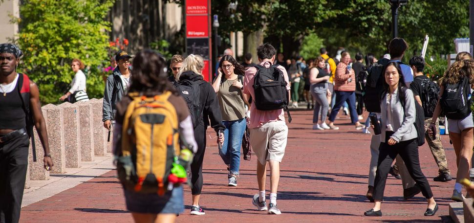 Photo of students walking up and down Comm Ave's brick sidewalk by Marsh Chapel. Many wear backpacks and it is a sunny day.