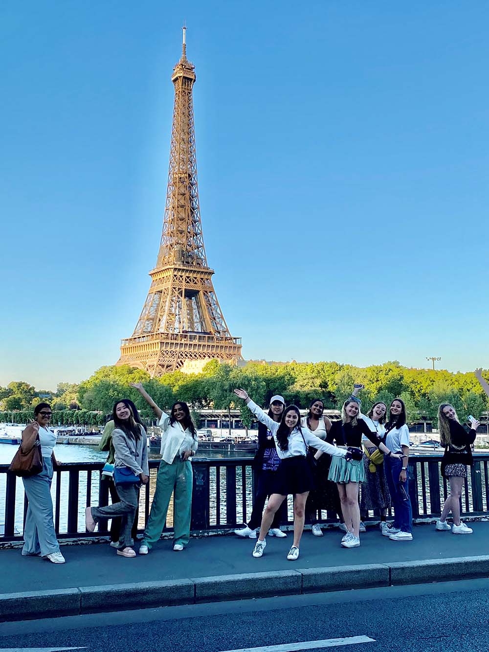 Photo of a group of students posing on a bridge with their arms pointed up towards the Eiffel tower behind them. The sky is bright blue. The students are a diverse group of young women.