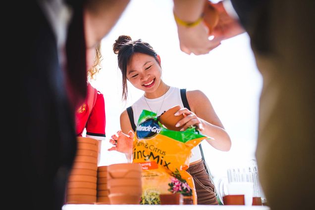 Photo of a group of students putting soil into small clay pots at a Weeks of Welcome event. At center, a young AAPI woman with black hair pulled back ads soil to a small clay pot. She smiles and wears a white shirt. The photo is taken at the level of the table, sort of looking up at the activity participants. Legs of our participants are seen blurred in the foreground.