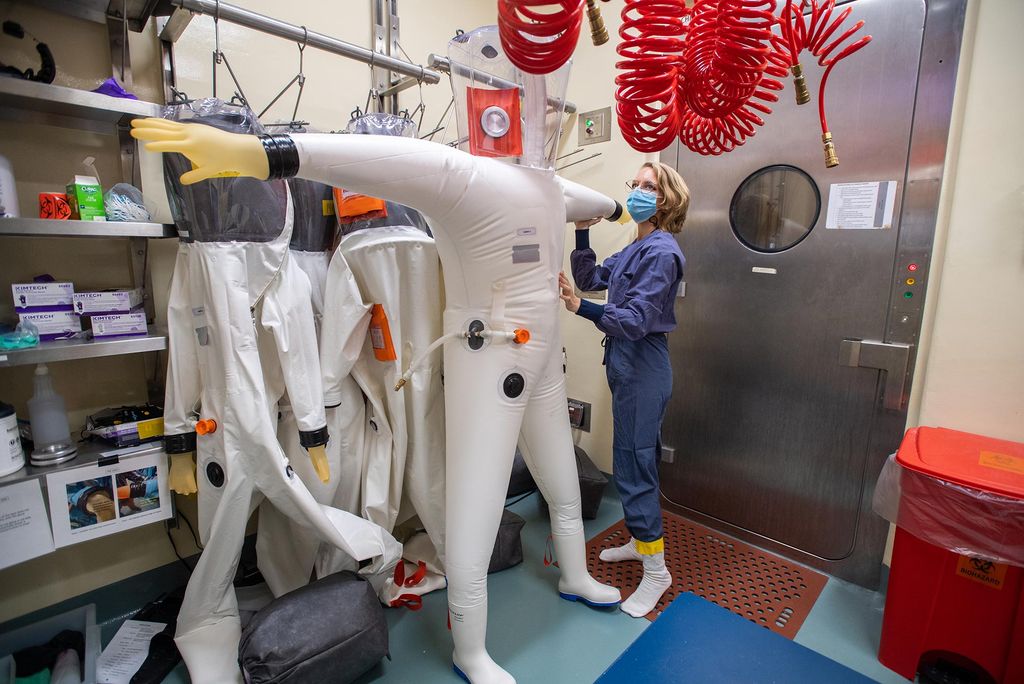 Photo of Pulmonary fellow Ellen Suder prepping her containment suit before working in the high containment lab, BSL-4, at the NEIDL September 1. She wears blue scrubs, a face mask, and socks. Other containment suits hang in the background and red curled tubes hang from the ceiling.