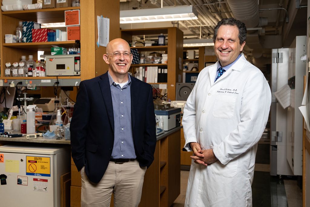 Photo of Joseph Mizgerd, left, and Darrell Kotton. Mizgerd wears a blue suit jacket, glasses, and smiles with his hands in his pockets. Kotton wears a white lab coat and smiles with his hands clasped in front of him. They stand in a lab, lots of vials are seen on shelves behind them.
