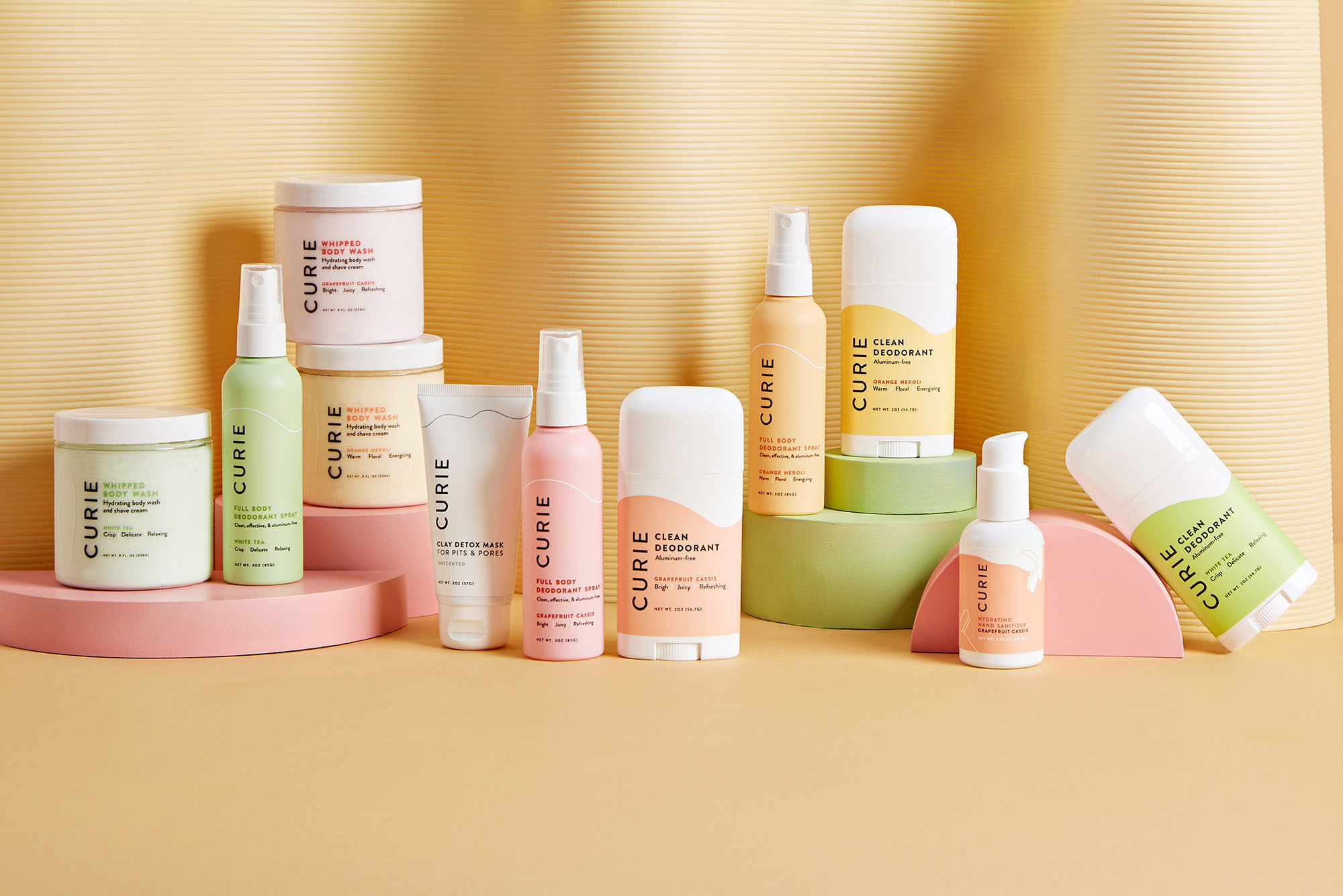 Aesthetic photo of an array of Curie products in various scents. Pastel-themed deodorants, lotions, and sprays are arranged in a light pastel yellow backdrop and floor.