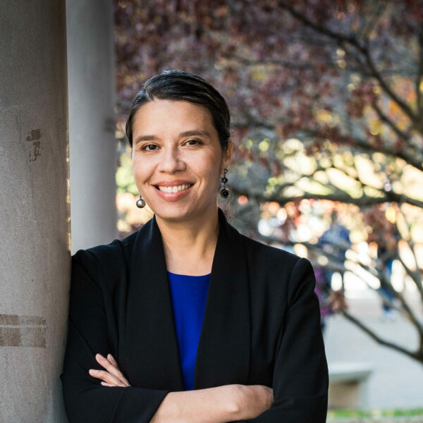 Headshot photo of Jasmine Gonzales Rose. A light-skinned black woman wearing a blue blouse and black blazer poses with arms crossed next to a tree.