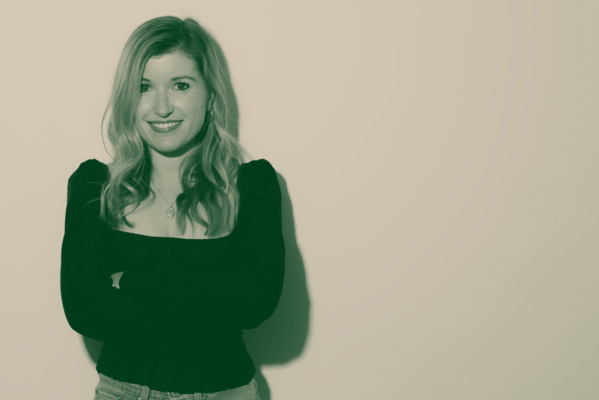 Photo: Studio headshot of Sarah Moret. A young white woman wearing a long-sleeved black shirt and jeans smiles in front of a white studio backdrop. Photo is overlaid with a greenish hued filter.