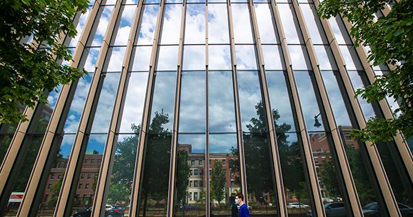Photo of Cloudy blue skies reflecting in the windows at the BU Kilachand Center. A long figure walks by on the sidewalk.