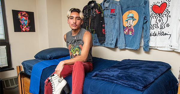 9/2/22 -- Boston, MA Matthew Garcia (Questrom’26) in his Claflin Hall dorm room Photo by Cydney Scott. Matt sits on a twin xl bed on risers with a blue bedspread. Holding a pair of white heels, Matt is also wearing red pants and a tank top with a fashionable graphic. Collared shirts of various designs hang on the wall behind.