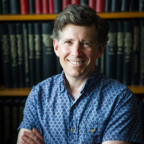 Photo: Headshot of Benjamin Wolozin posing and standing in front of bookcase with arms crossed. A white man wearing a blue collared shirt smiles.