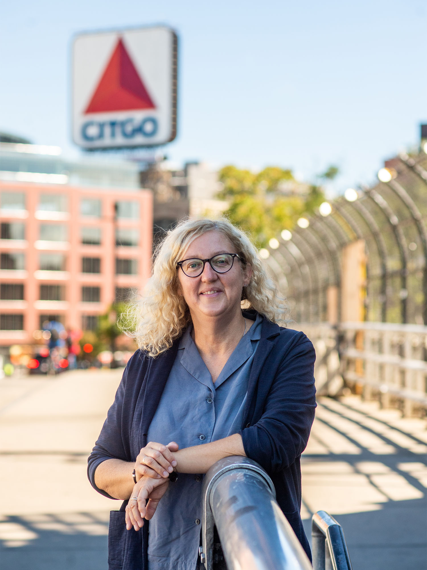 Photo: Loretta Lees, a white woman with curly blonde hair and wearing glasses, a blue collared shirt, and navy blue cardigan, smiles and poses against a railing outside. A bridge and giant Citgo sign can be seen in the blurred background behind her).