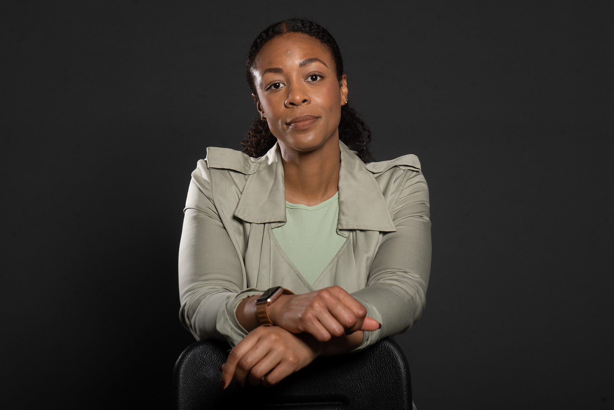 Studio portrait of Amanda Bailey. A black woman with straight black hair and a light smile leans on a chair with hands intertwined on top of a black chair. She wears a black and white blouse and no-frame glasses.