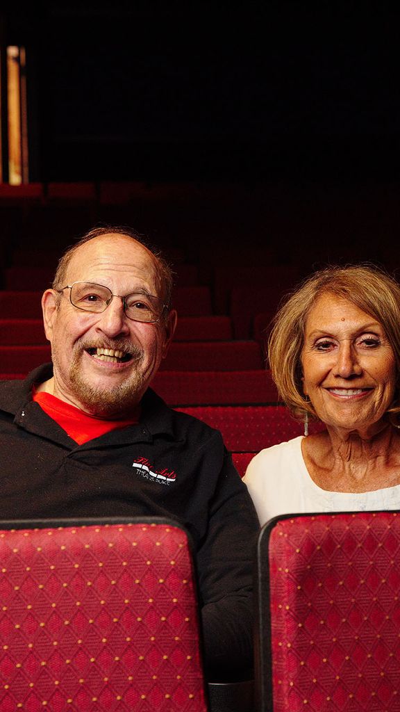 Photo: Alums Les Gordon (left) and Dafna Krouk-Gordon sit in an empty theater and pose for the camera. An older white man wearing glasses and a black long sleeved shirt sits tot he left of an older white woman wearing a white top. They both sit in an empty theater with red seats and smile for the camera.