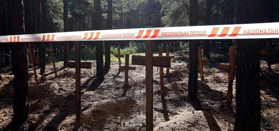 Photo: Wooden crosses mark the graves of Izium residents killed by Russian occupiers during exhumation procedures at a mass burial site in Izium that was liberated by the Ukrainian Armed Forces from Russian troops, Kharkiv Region, northeastern Ukraine. Numerous mounds of dirt are marked by crosses in a wooded area. A tape is pulled across the scene, with Ukrainian writing on it.