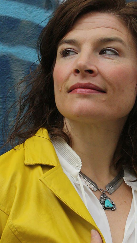 Photo: Jenn Lindsay poses for a photo. A white woman with wavy brown hair and wearing a white collared shirt and yellow jacket poses in front of a blue wall and looks to the left.
