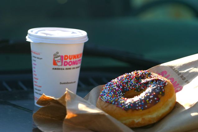 Photo: A Dunkin' donuts branded paper cup of coffee and a chocolate frosted donut with rainbow sprinkles lies on a Dunkin' Donuts brown paper bag is shown on a dashboard.
