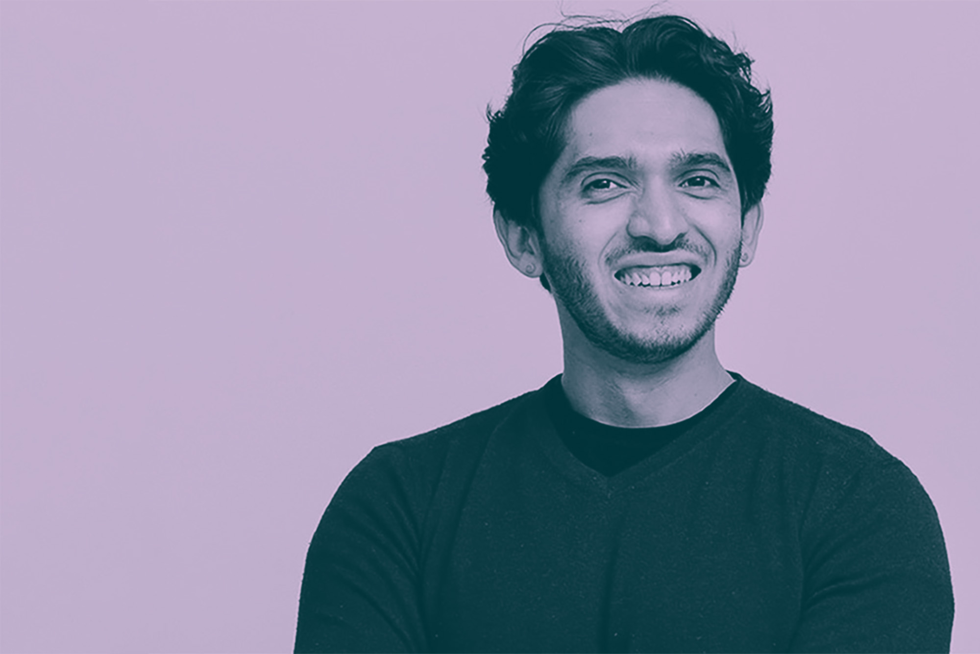 Photo: Headshot of Juan Ramirez, a Latinx man with black hair and wearing a black shirt and burgundy sweater, smiles and poses with arms crossed. Photo is overlaid with a lavender hued filter.