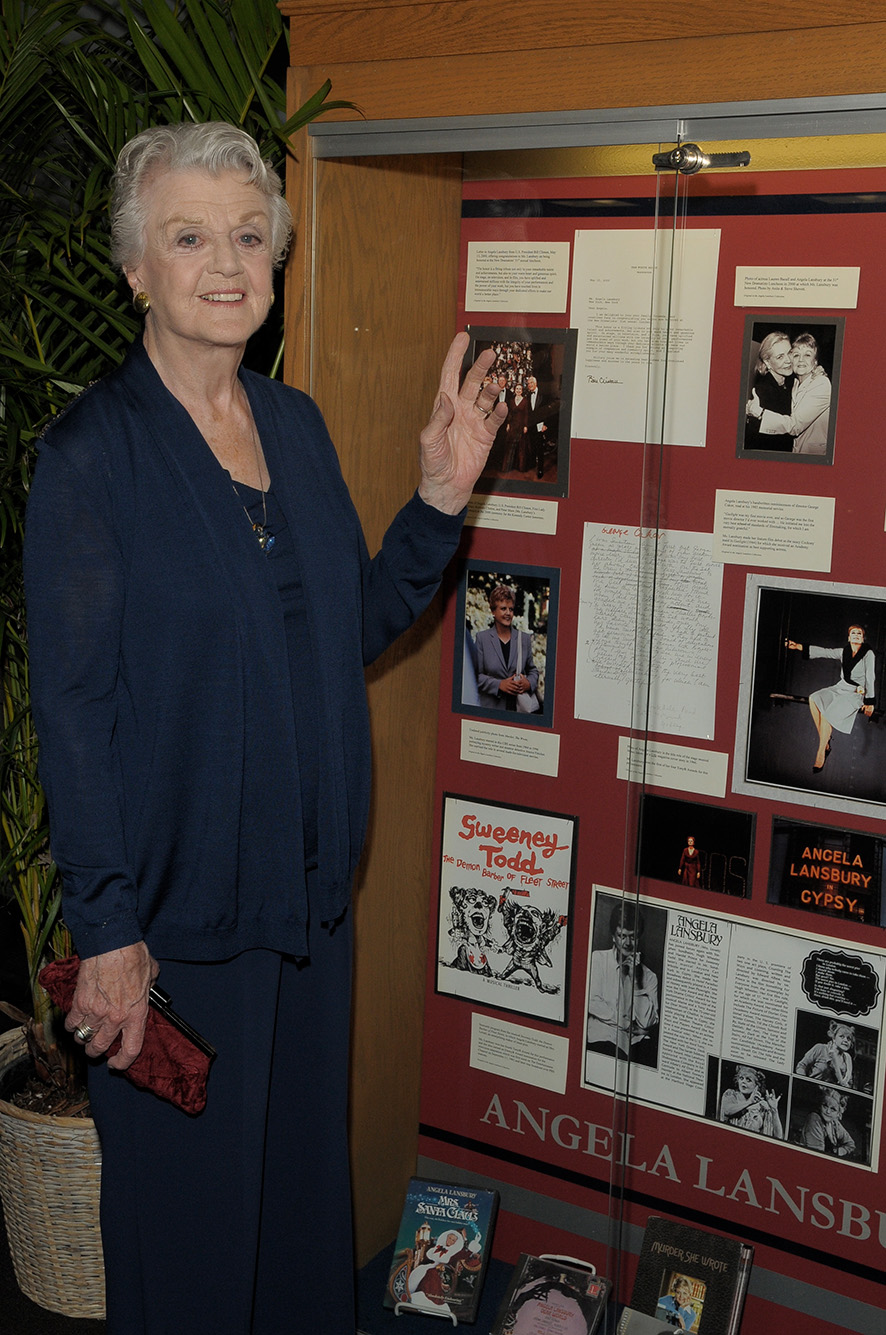 Photo: Lansbury in 2011 in front of a case of memorabilia from her career on view at the Howard Gotlieb Archival Research Center. An older white woman with graying hair poses and points to a large glass case featuring various works.