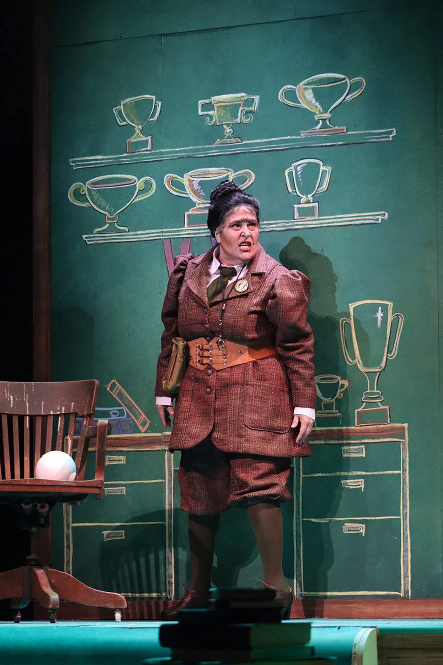 Photo ofJohanna Carlisle-Zepeda as the evil Miss Trunchbull. She wears a steampunk-style brown trenchcoat and pants and looks menacing as she sings in front of a fake chalkboard.