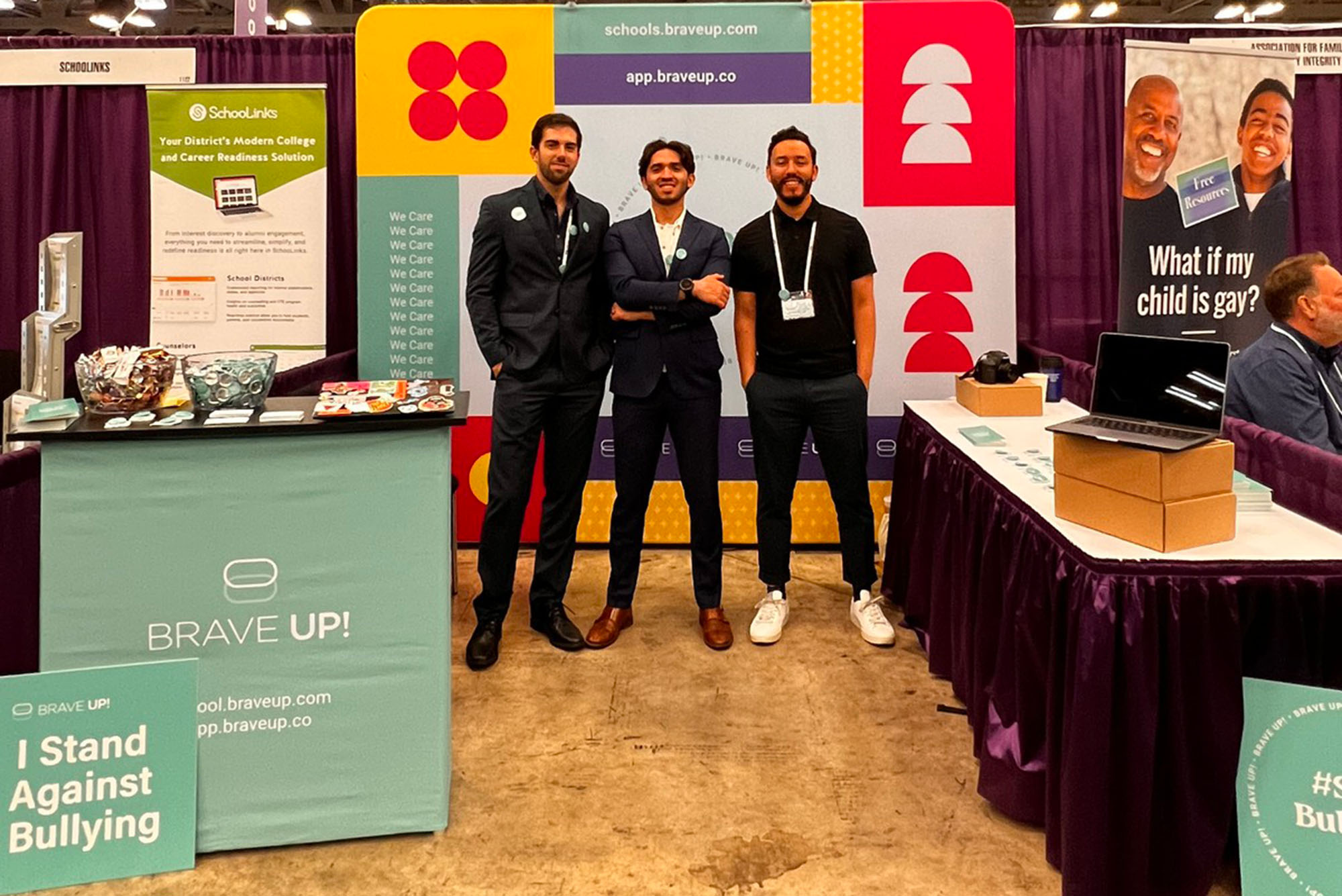 Photo: Ramirez (center) with Brave Up! higher-ups Enrique De Lima (CAS’21) (left), head of growth, and Álvaro Carrasco Arriaza, cofounder and CEO, at a convention for school counselors in New York City. 3 men wearing black suit ensembles pose near a BraveUp themed booth in a conference hall.