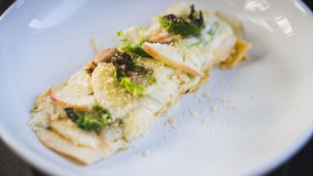 Photo: A large piece of butternut squash manicotti topped with Brussels sprouts, pickled apple, and almonds is shown on a white plate. A slight sprinkling of Parmesan cheese tops the dish.