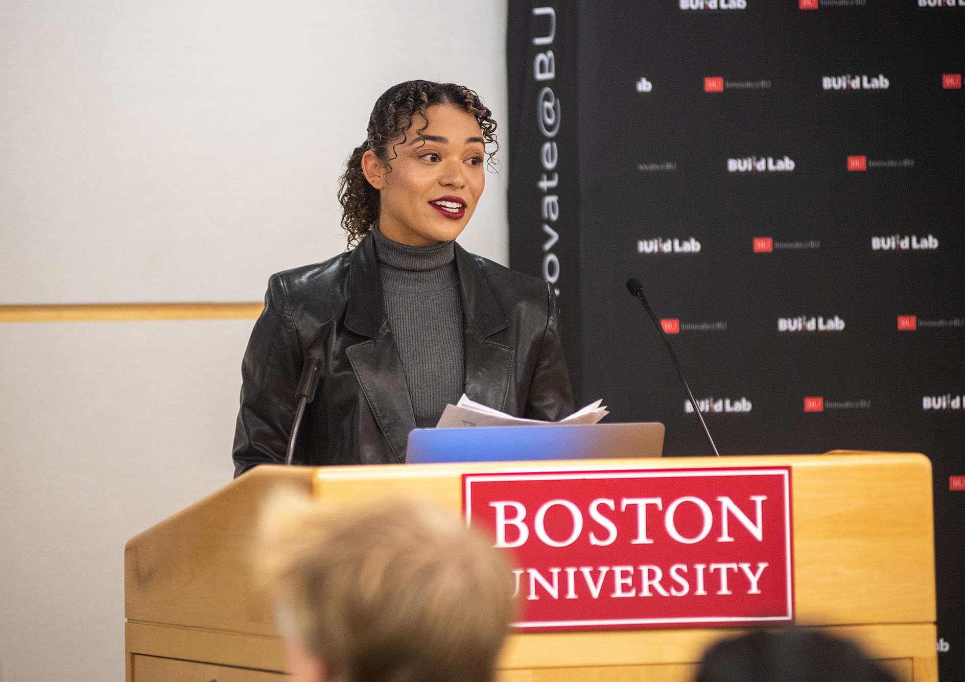 Photo: Isabelle Bryan, a young tan woman wearing a grey turtleneck and leather blazer, speaks into a microphone at a podium with a sign that reads "Boston University" on the front of it.