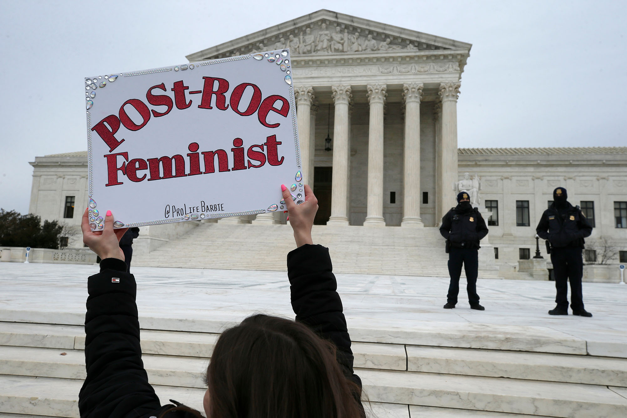 Photo of a protestor holding a sign that reads "Post-Roe Feminist" in front of the exterior of US Supreme Court building. She holds up sign as two officers look on from the distance.