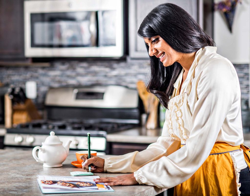 Photo of Priya Shah, wearing a white ruffly blouse and a mustard-colored apron, writing out a shopping list as she reads a page of LVNGbook. She stands at a marble kitchen island in a sleek modern kitchen.