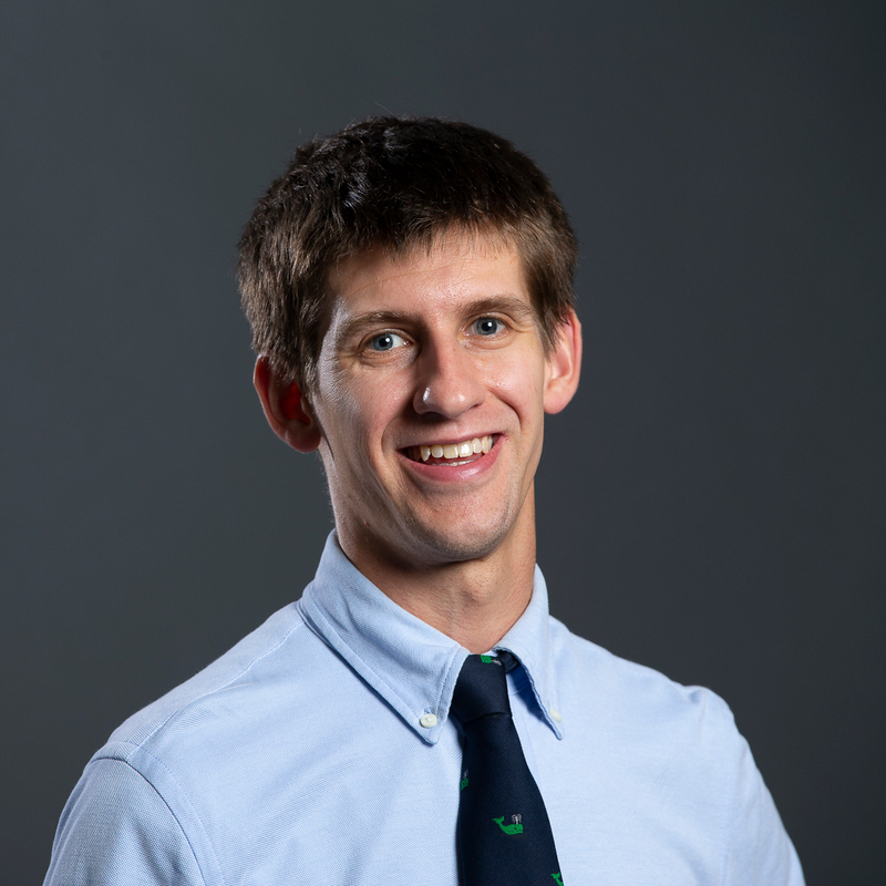 Photo: Posed headshot of Thomas Byrne. A white man with straight brown hair and wearing a light blue collared shirt and black tie smiles and poses in front of a dark grey backdrop.