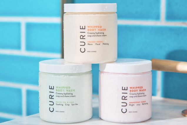 Photo: Display of three bath scrubs on a marble bathroom countertop. They are arranged in a mini pyramid with branding for the brand Marie.