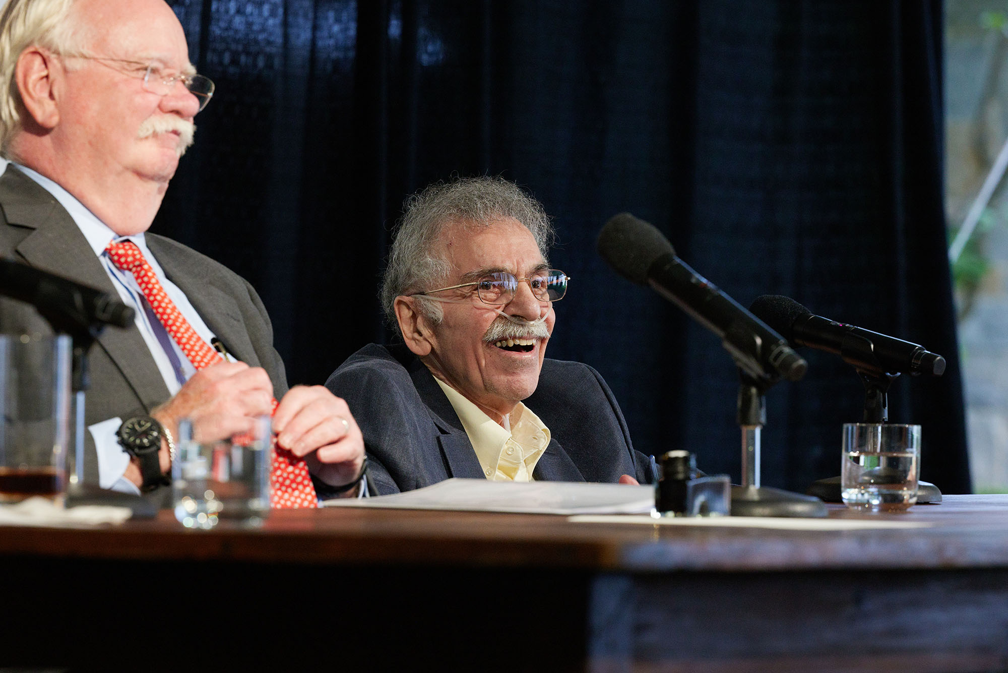 Photo of President Brown and Edward Avedisian seated at a large, dark brown table one stage, smiling and looking off to the right at the crowd.
