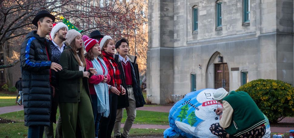 Sam Weinberger (CFA’21) paints the BU rock with holiday flair as the student a cappella group the Allegrettos watches and sings on. A group of students wearing holiday hats and scarves sing as a young man bends down to spray paint a giant rock with a snowman holiday scene.
