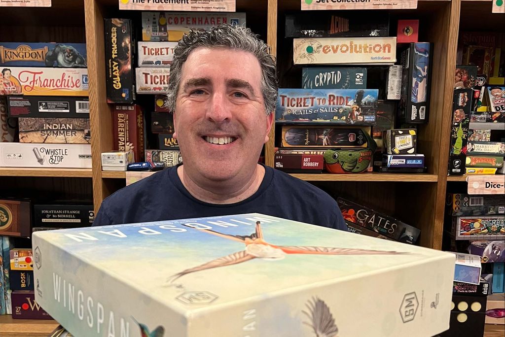 Photo: Neil Goldberg, a white man with dark silver gray hair, smiles as he sits and looks over a large stack of board games. Behind him, numerous wooden shelves filled with more board games are also shown.