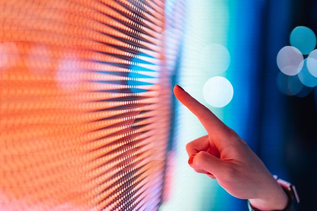 An outstretched finger pointing towards a digital screen lit up with oranges, reds, and blues