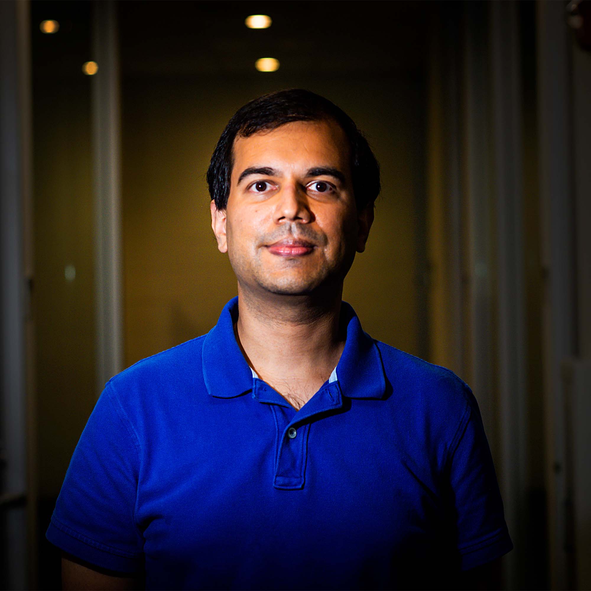 A portrait of Mayank Varia on a dark background, wearing a blue shirt. He looks pensively toward the camera.