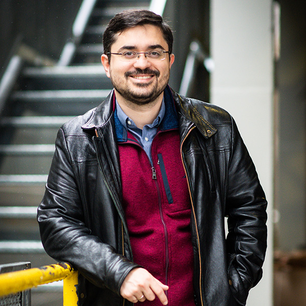 Photo: Manos Athanassoulis, Assistant Professor of Computer Science, stands and leans against a yellow railing as he poses for a photo. A tan man wearing wire frame glasses, a red sweater vest, and leather jacket, smiles and poses for the photo. A set of blurry photos can be seen behind him.