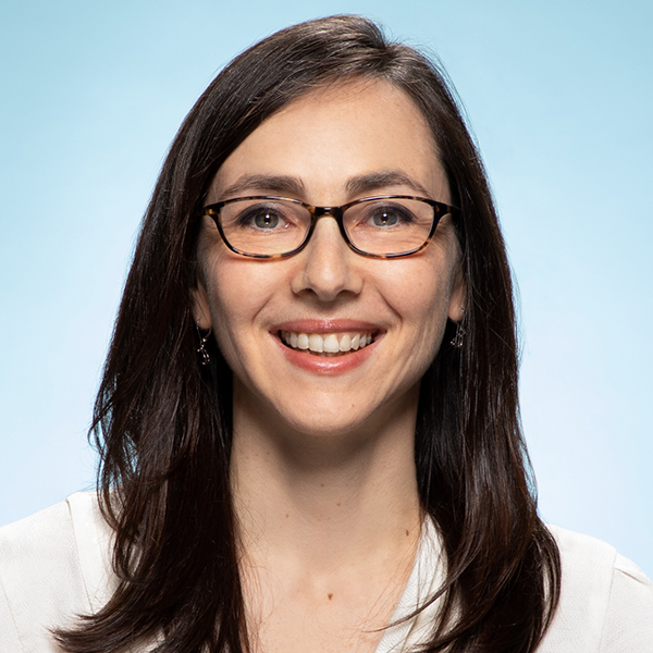 Photo: Headshot of Masha Kamenetska. A smiling white woman with long brown hair wearing a white blouse and glasses, smiles and poses in front of a light blue background.