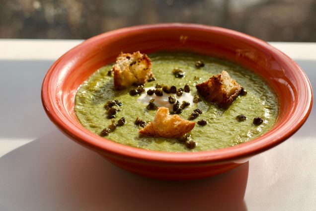 Photo: a red bowl sat next to a window letting in natural light is filled with a dark green soup and topped with homemade croutons, a dollop of cheese, and capers.