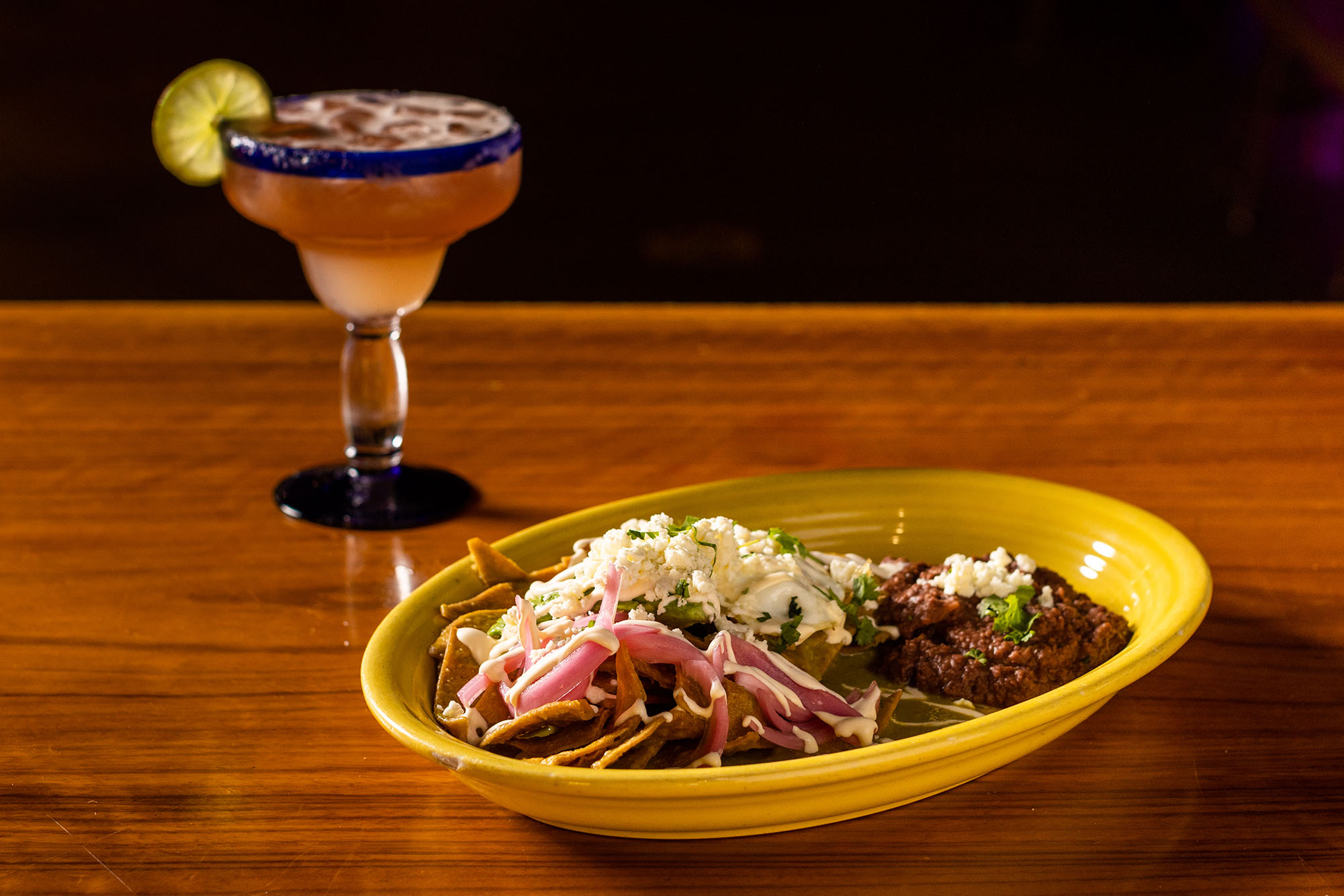 Photo: A plate of Beef Birria, nachos with shredded cheese, and a classic margarita are displayed on wooden table in front of a black backdrop.
