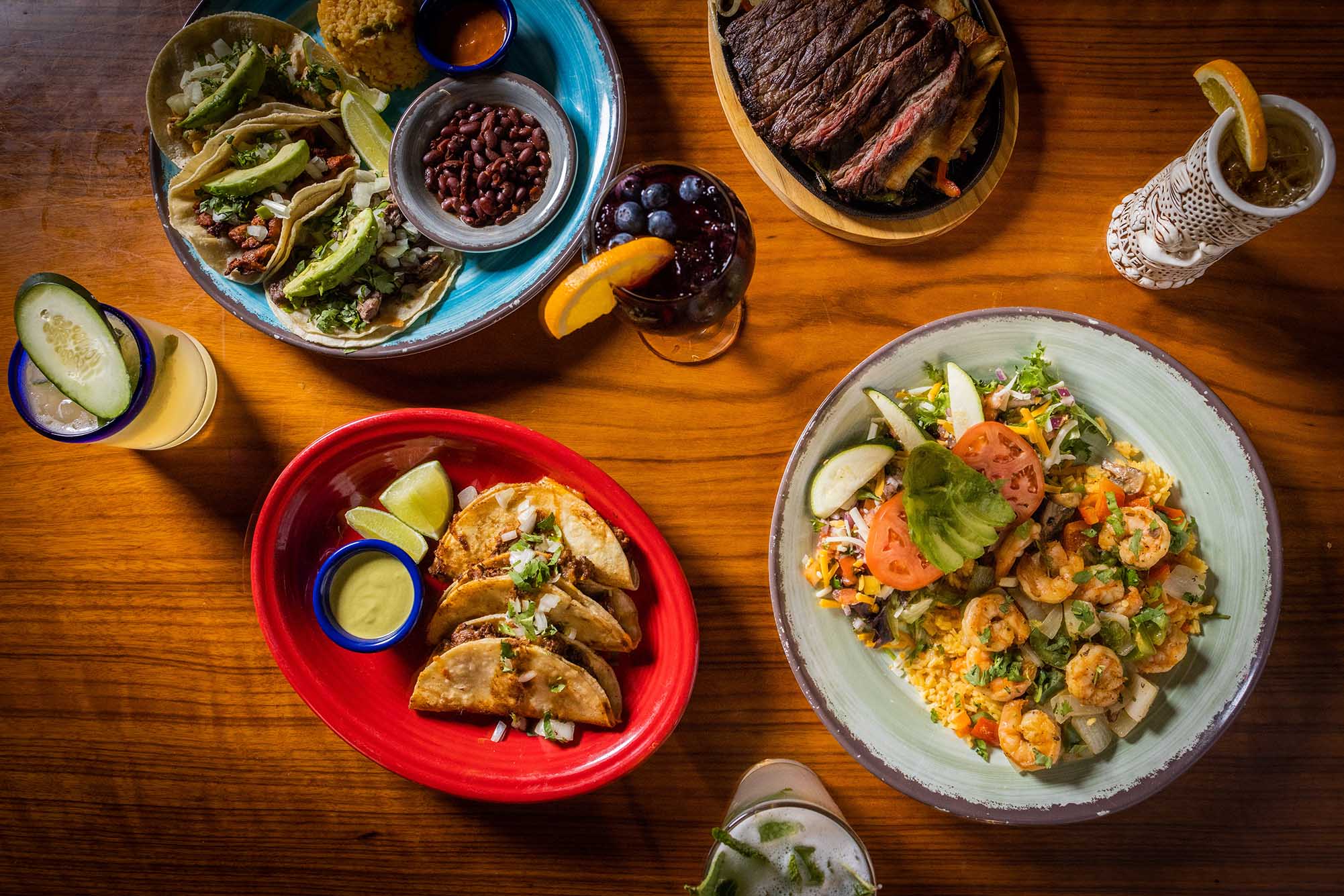 Photo: Overhead shot of an array of food dishes including but not limited to various styles of tacos, carne asada, and shrimp are displayed. A few fruity alcoholic drinks, including a margarita, are also shown among the dishes. They're all displayed on a wooden table.