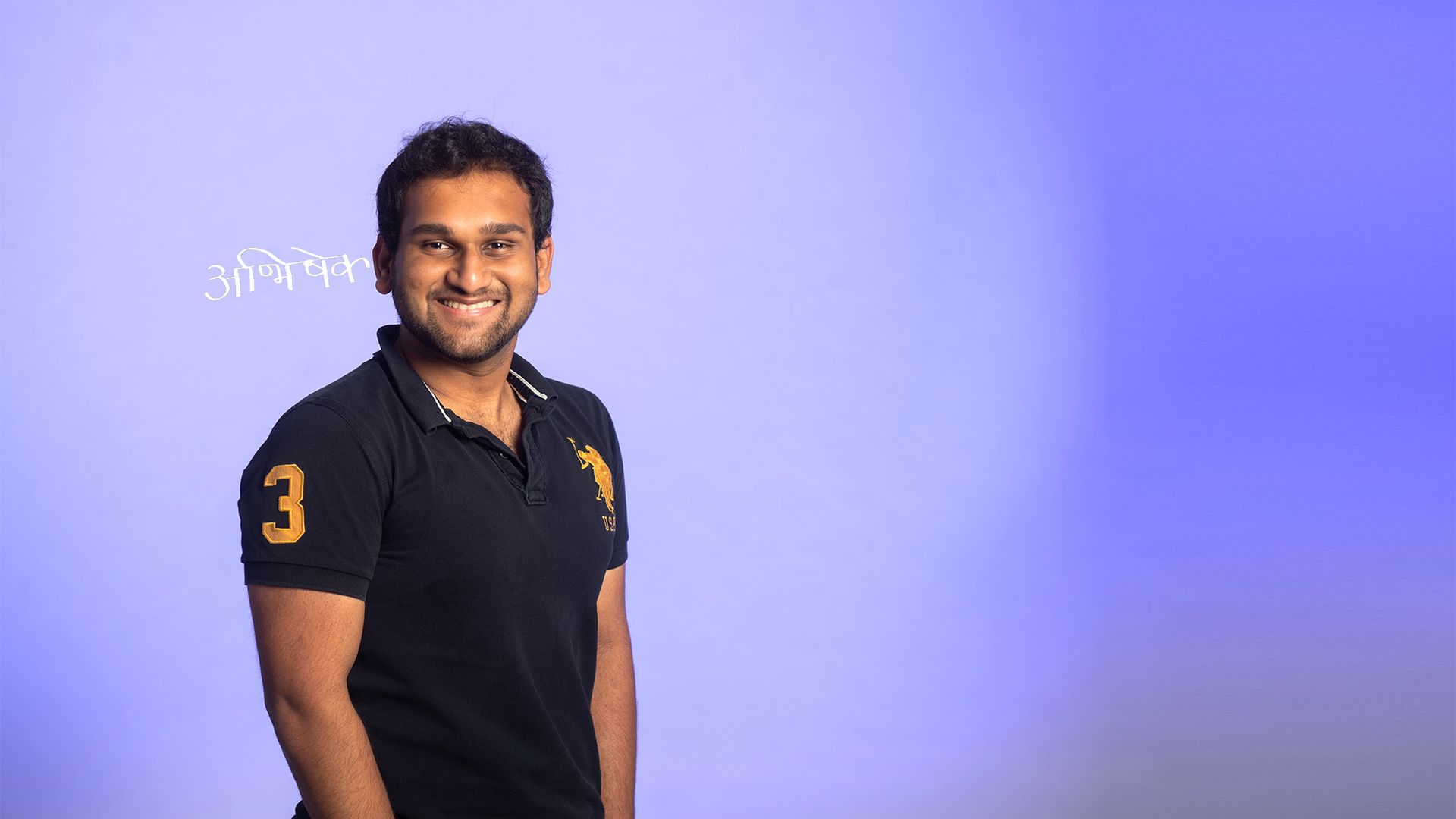 Photo: A West Asian man with a black short sleeve and his hands in his pockets. He is smiling into the photo.