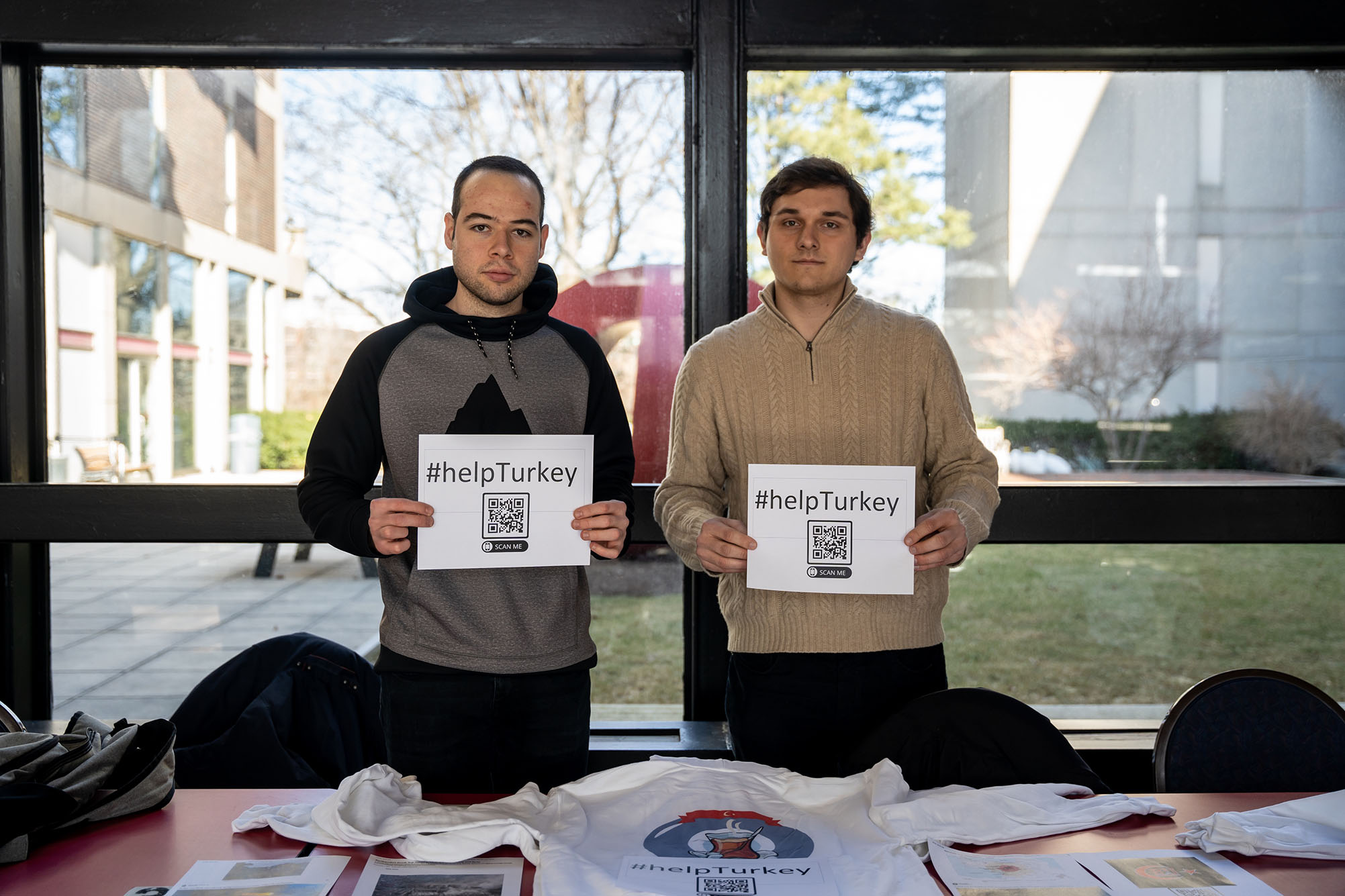 Photo: Derya Benal (ENG'23), left. and Kevin Clenard (CAS, Pardee '24) represent the Turkish Student Association who are selling sweatshirts for $30 to raise money for relief aid in Turkey. Two Turkish students stand in front of a large window and hold up a signs that read "#helpTurkey" and a QR code that leads to the GoFundMe page. The table in front of them shows shirts and sweatshirts that also read "#helpturkey" as well as information on the earthquake.