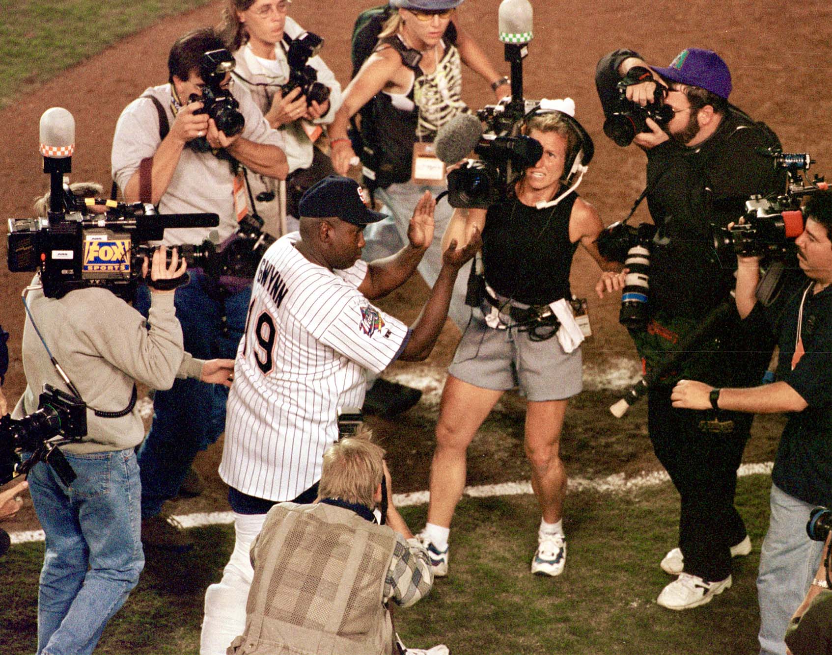 Photo: Camera operator Deena Sheldon (COM’84) (center) at the 1998 World Series. A white woman carrying a large camera pushes to the front of a group of others holding large film and photo cameras to get a shot of a baseball player on the field.