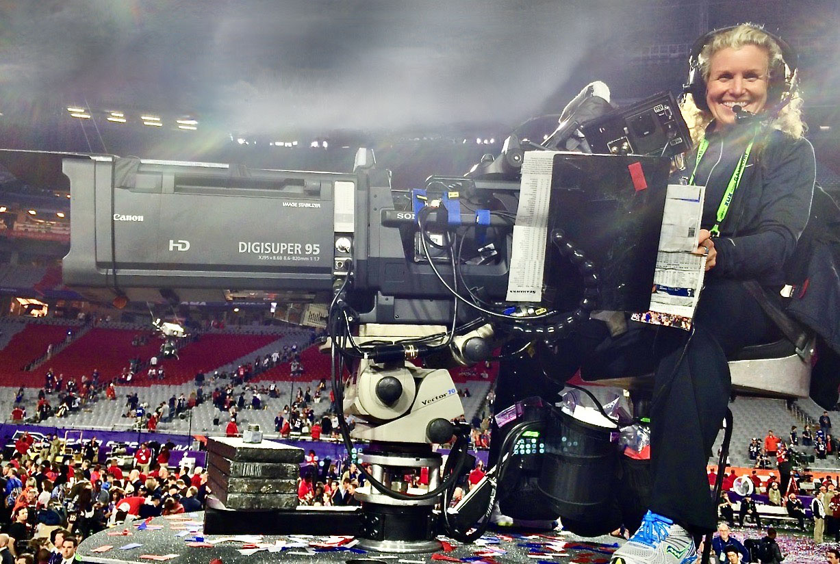 Photo: At Super Bowl XLIX in 2015, New England Patriots vs the Seattle Seahawks, in Glendale, Ariz. A white woman with long, curly, dirty blonde hair smiles as she sits behind an large film camera in an emptying football stadium.