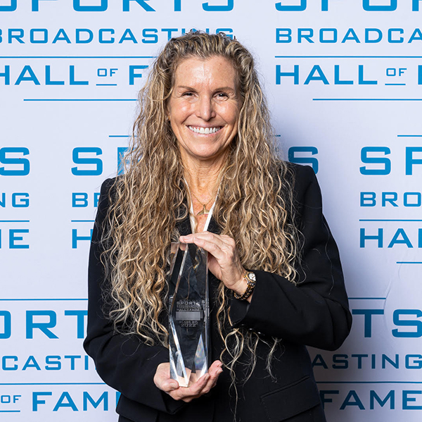 Photo: Sheldon at the Sports Broadcasting Hall of Fame ceremony in December 2022. A white woman with long, curly, dirty blonde hair and wearing a black blazer and pants smiles as she holds up a glass trophy. The backdrop behind her reads "Sports Broadcasting Hall of Fame".