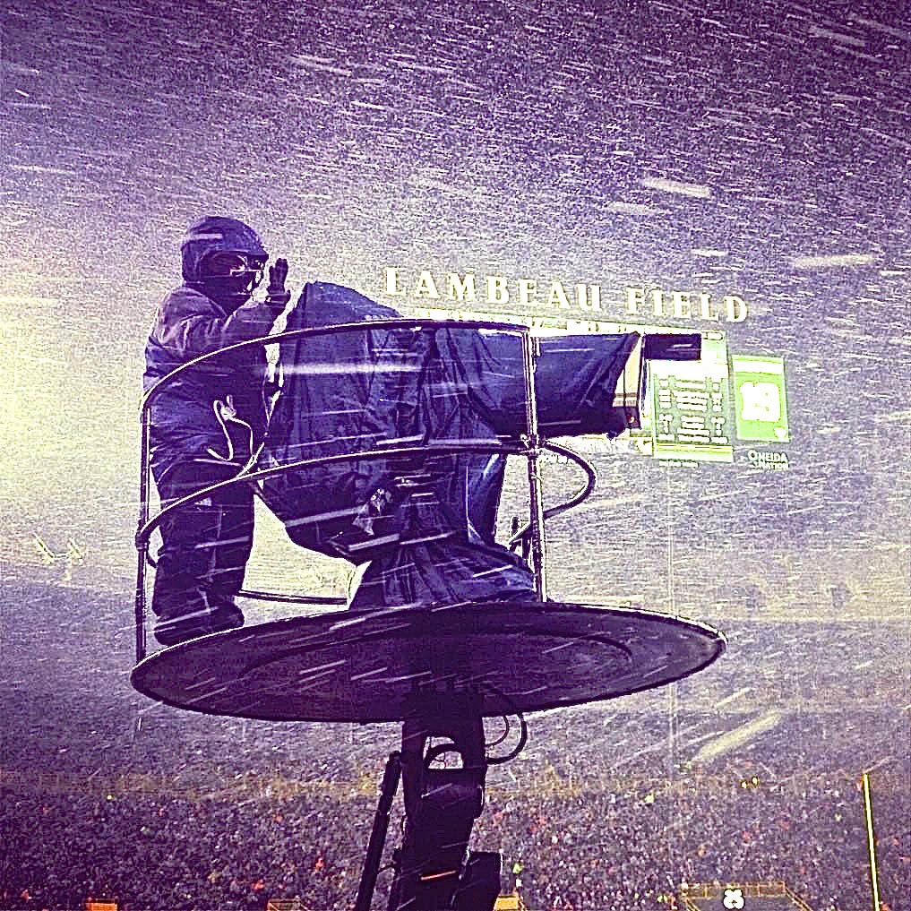 Photo: Lambeau Field, home of the Green Bay Packers, in 2012. A camera operator is shown bundled up in a black coat as they stand on a platform with a large film camera that is covered in a large black tarp. Around them snow billows into the night and the scene is highlighted by the stadium lights.