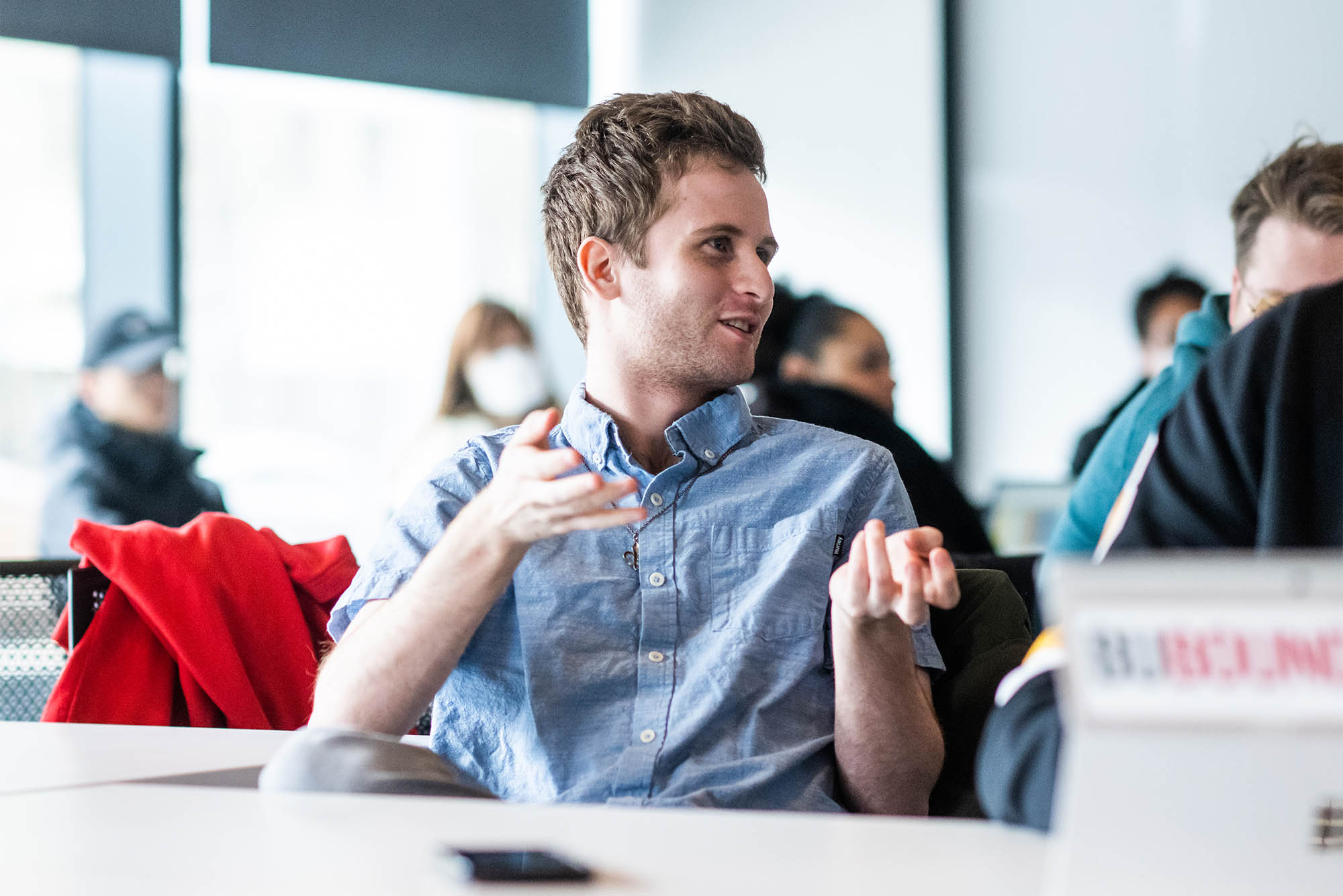 Photo: A white man wearing a light blue collared shirt gesticulates as he talks to someone on his right. Other students can be seen in the blurred background.