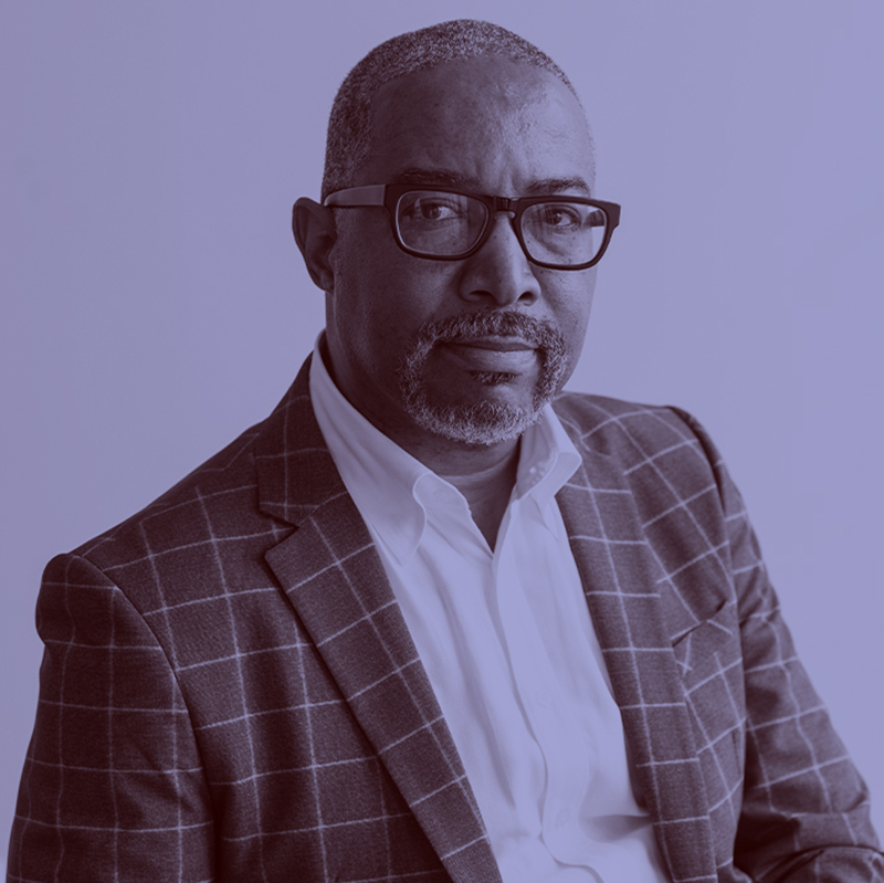 Photo: Frank Rudy Cooper, a Black man with greying shaved hair and beard and wearing a white collared shirt and teal jacket, poses for a photo. A light purple filter overlay is placed over the photo.