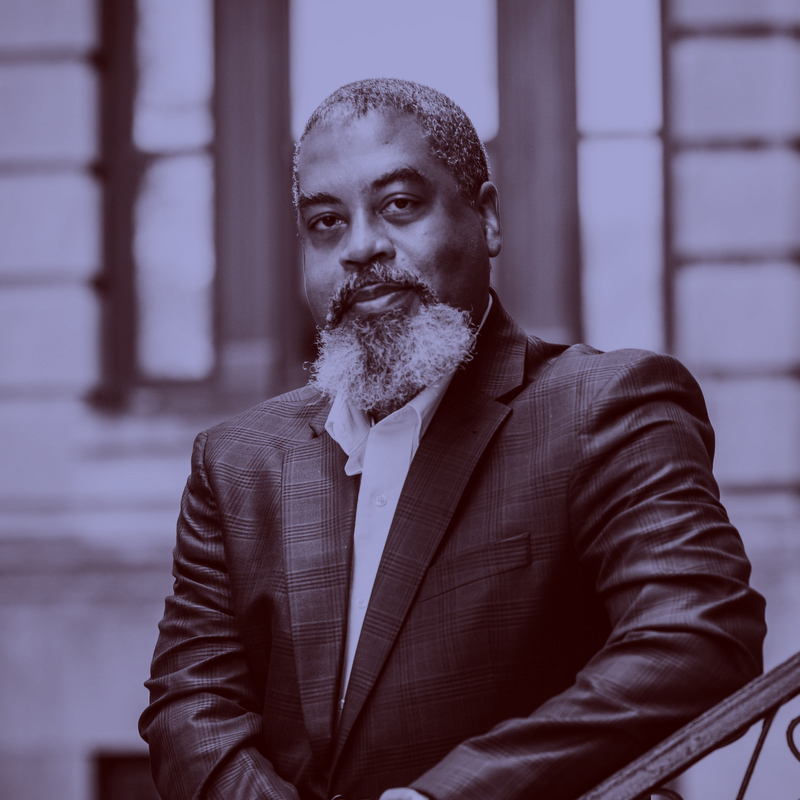 Photo: Phillipe Copeland, a Black man with a long, grey beard wearing a white collared shirt and black blazer, sits and poses on an outdoor bench. A light purple filter overlay is placed over the photo. 