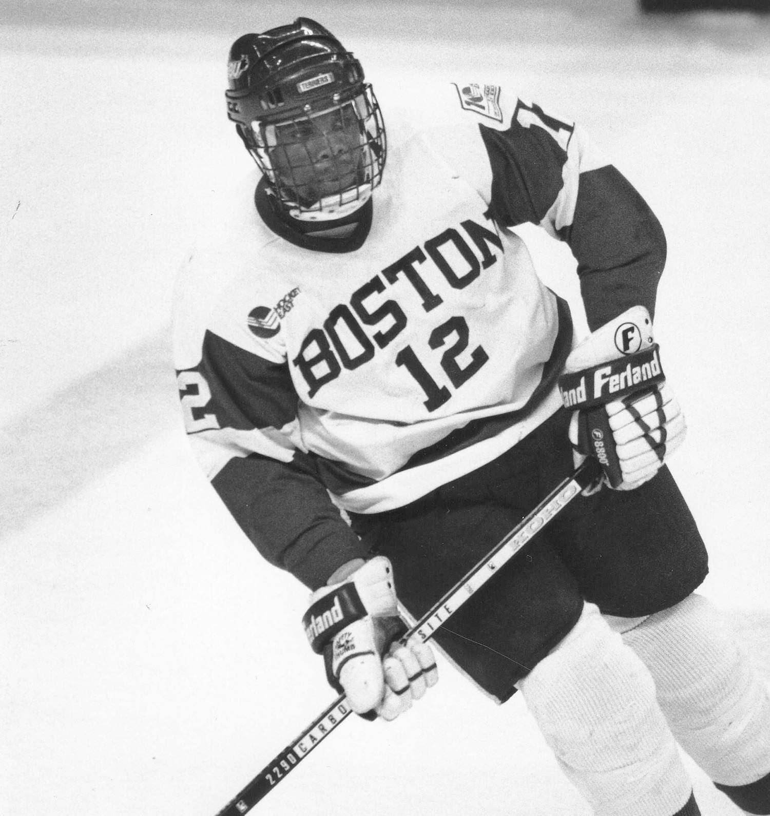 Photo: Black and white photo of Mike Grier, a Black man wearing a BU "Boston" ice hickey uniform and helmet, as he holds his stick and skated during a game.