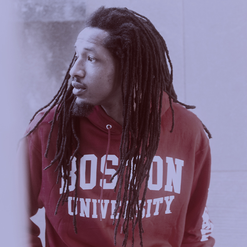 Photo: Darien Pollock, a Black man with long dreadlocs and wearing a "Boston University" hoodie, smiles and poses for the photo. A light purple filter overlay is placed over the photo.