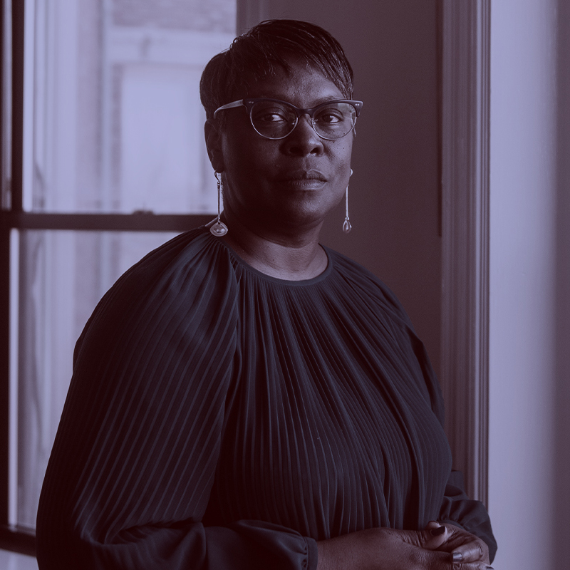 Photo: Margarita Guillory, a Black woman with short black hair wearing glasses and a dark blue blouse, stands with hands folded as she poses for a photo. A light purple filter overlay is placed over the photo.